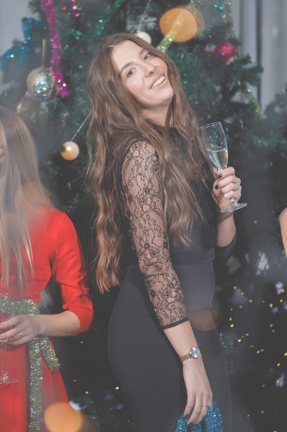 Hermosa mujer divirtiéndose con una copa de champán cerca de un árbol de Navidad. Una mujer ríe, sonríe, posa. Filtro de grano y ruido vintage especial, luces borrosas.