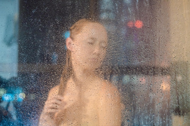 Hermosa mujer en la ducha detrás de un vidrio con gotas en el fondo de una ventana con una vista panorámica de la ciudad de noche