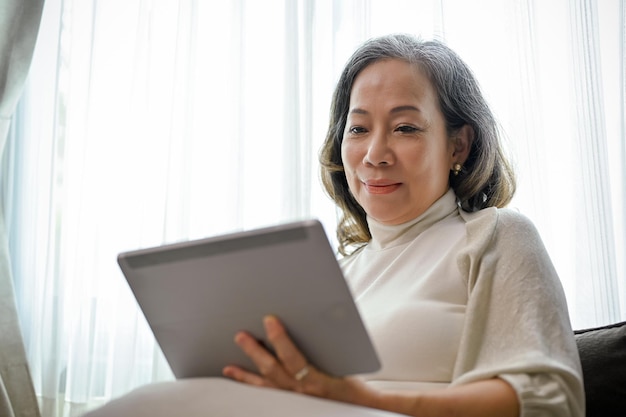 Una hermosa mujer de edad asiática ve un videoclip en línea en una tableta digital en su sala de estar