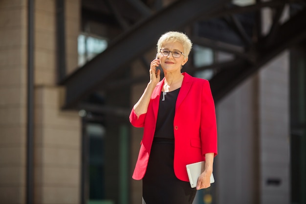 Hermosa mujer de edad avanzada con un corte de pelo corto y gafas en una chaqueta roja con un teléfono