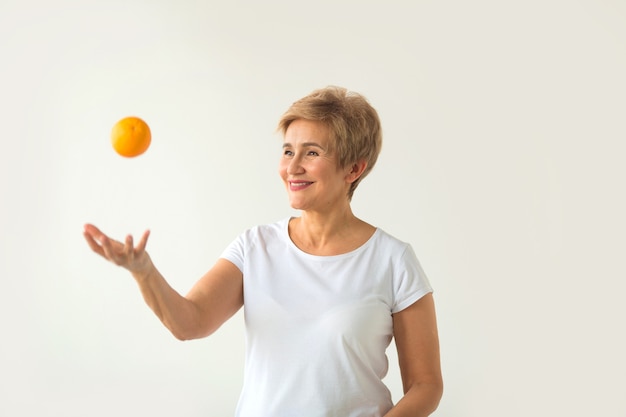 Hermosa mujer de edad, una camiseta blanca arroja una naranja sobre un fondo blanco.