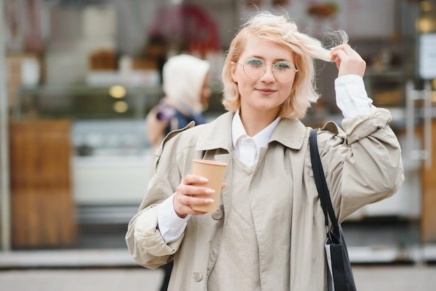 Hermosa mujer elegante con buena ropa de moda caminando en la calle y tomando un café en la taza para llevar con buen humor de verano cerca de la cafetería de la calle.