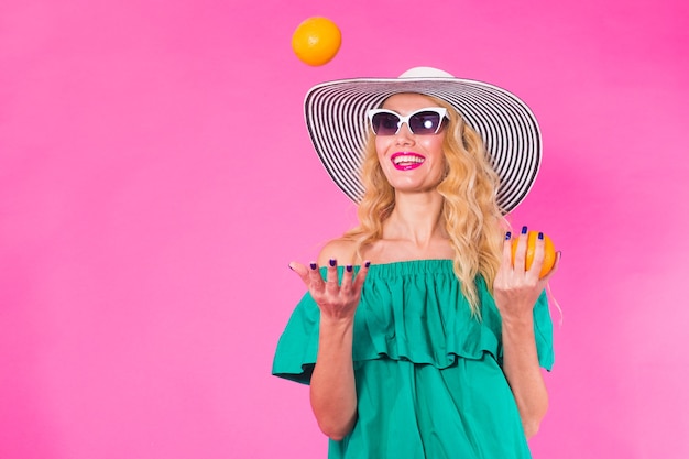 Hermosa mujer elegante en gafas de sol y sombrero con naranjas divirtiéndose sobre pared rosa. Concepto de verano, vacaciones y moda.