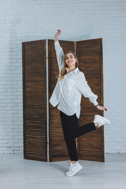 Una hermosa mujer elegante lleva una camisa blanca de algodón y pantalones negros. Alegre chica emocional con zapatillas blancas.