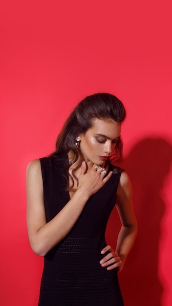 Hermosa mujer elegante con maquillaje, lápiz labial rojo en elegante vestido negro, lleva joyas, sobre fondo rojo.