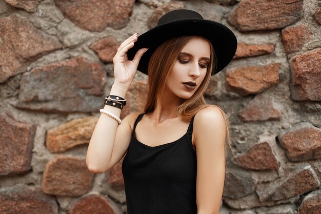 Hermosa mujer elegante en ropa negra de moda con sombrero cerca de un muro de piedra