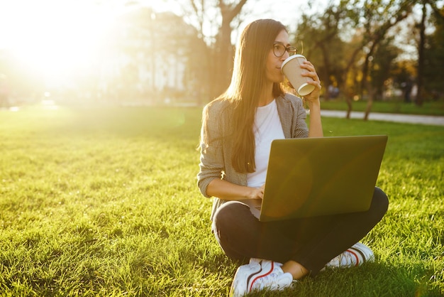 Hermosa mujer elegante sentada en la hierba verde con una computadora portátil y un teléfono en las manos Concepto de estilo de vida