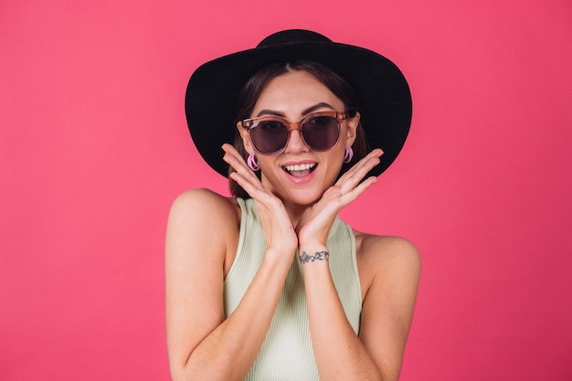 Hermosa mujer elegante con sombrero y gafas de sol posando sobre pared roja rosa