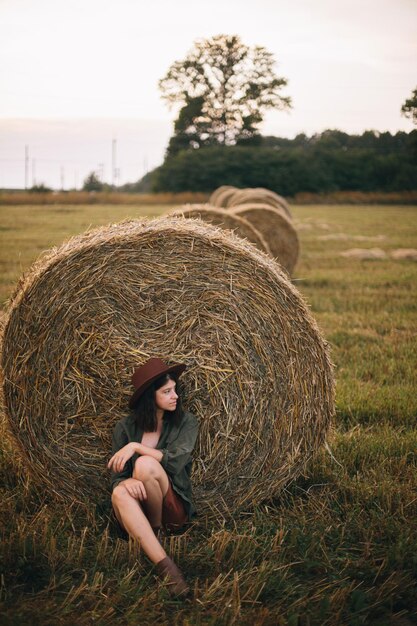 Hermosa mujer elegante con sombrero sentada en pacas de heno en el campo de la noche de verano Relajándose en el campo