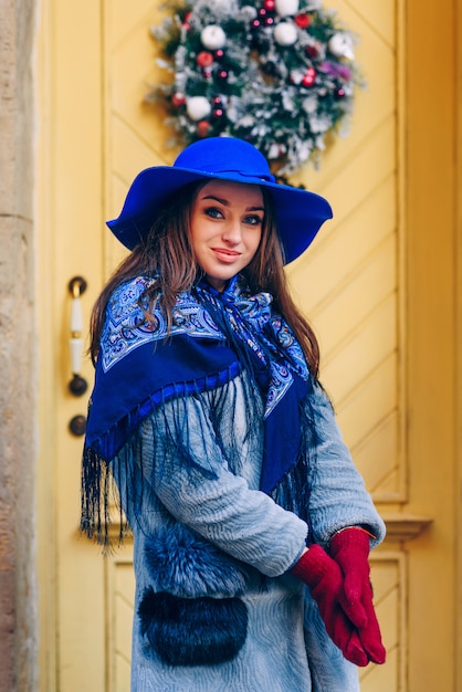 Hermosa mujer elegante con una sonrisa perfecta con sombrero azul y bufanda caminando por la calle. Mujer cerca de la puerta amarilla con adornos navideños e inscripción Bienvenido.