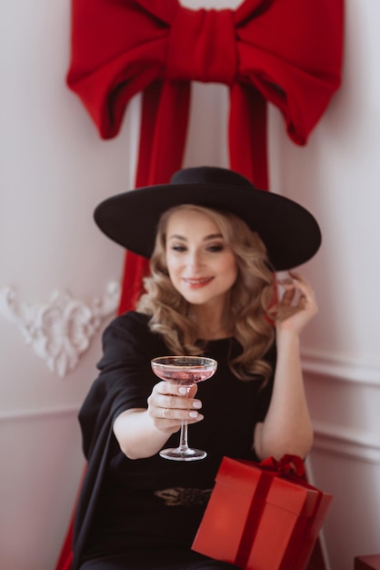 Hermosa mujer elegante con vestido festivo negro y sombrero con copa de champán y regalos de Año Nuevo Enfoque selectivo suave