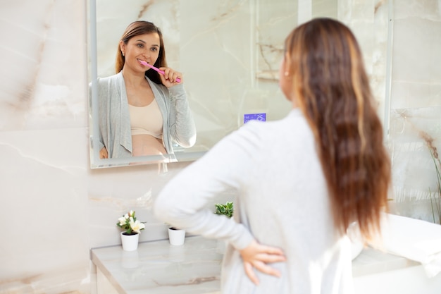 Una hermosa mujer embarazada feliz en ropa de casa se cepilla los dientes frente a un espejo en un baño luminoso. Estilo de vida. Rutina matutina y vespertina. Higiene. Cuidado de la salud. Foto de alta calidad