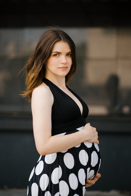 Una hermosa mujer embarazada joven con estilo en un vestido negro con lunares blancos sostiene sus manos sobre su estómago. Feliz esperando al bebé