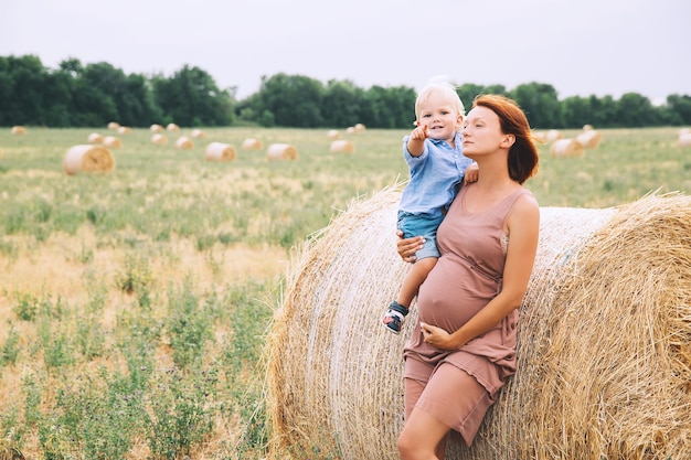 Hermosa mujer embarazada y niño divirtiéndose en el campo de trigo con montones de heno en el día de verano en la naturaleza