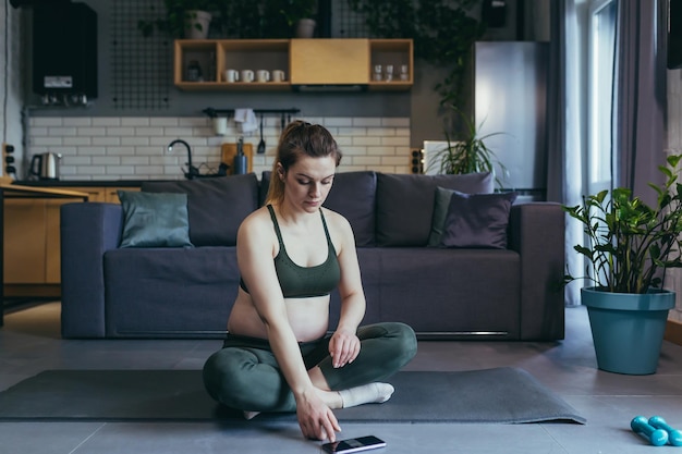 Hermosa mujer embarazada practica deportes en casa en línea se sienta con el teléfono en una colchoneta deportiva con un uniforme deportivo hace ejercicios está satisfecha