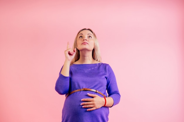 Hermosa mujer embarazada rubia en un vestido con el dedo del vientre (panza) apuntando hacia arriba en el estudio sobre un fondo rosa copyspace.