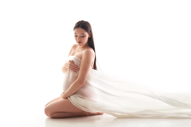 Hermosa mujer embarazada sentada en el suelo de seda blanca