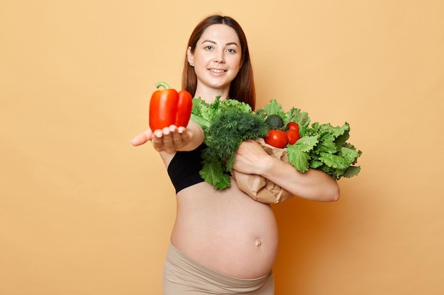 Hermosa mujer embarazada sonriente abraza verduras frescas posando aisladas sobre fondo beige futura madre con el vientre desnudo mostrando pimienta orgánica