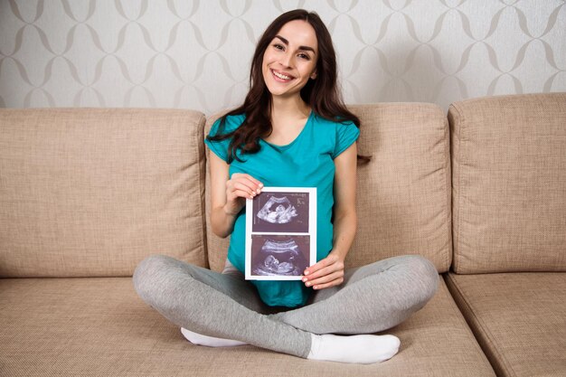Hermosa mujer embarazada sonriente sentada en el sofá sosteniendo una imagen de ultrasonido y mostrándola a la cámara Esperando al niño