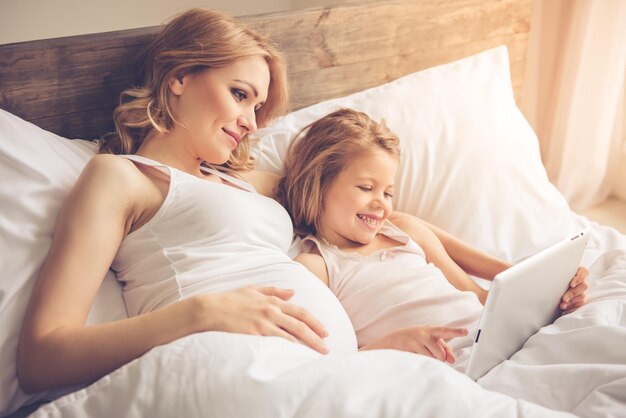 Hermosa mujer embarazada y su linda hijita están usando una tableta digital y sonriendo mientras pasan tiempo juntos en la cama