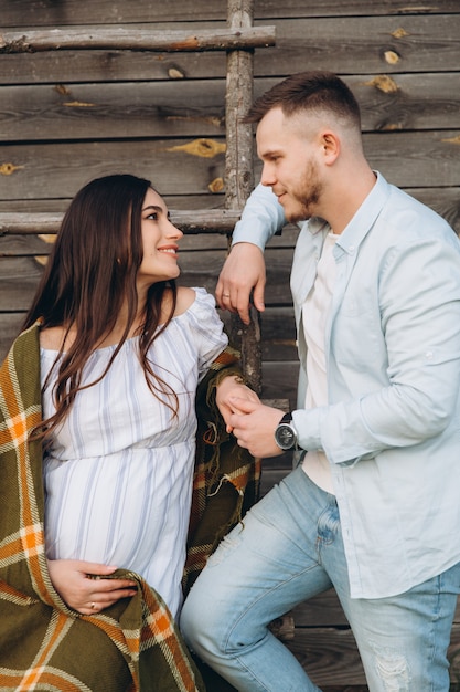 Hermosa mujer embarazada y su marido posando cerca del edificio de madera. Elegante pareja feliz se aman. Moda y estilo rústico.