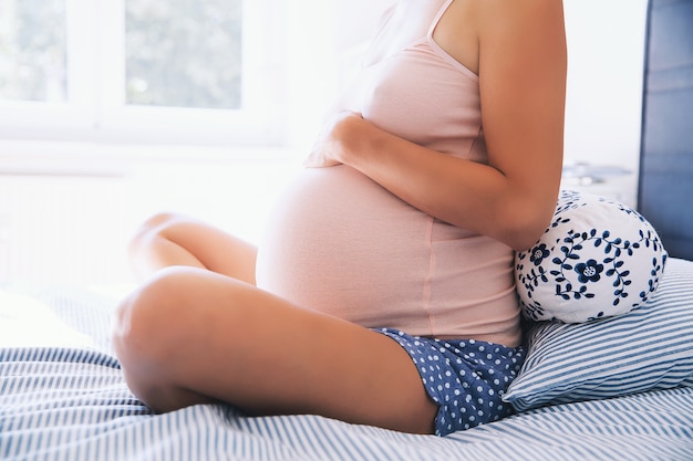 Hermosa mujer embarazada tiene las manos sobre el vientre en el dormitorio en casa Joven madre esperando de un bebé