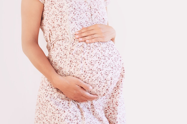 Hermosa mujer embarazada vestida tiene las manos en el vientre sobre fondo blanco.