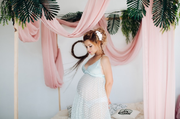 Hermosa mujer embarazada en vestido camisón posando dentro de la decoración con flores.