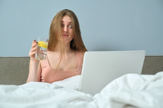 Hermosa mujer emocional con la boca abierta sentada en la cama con ordenador portátil