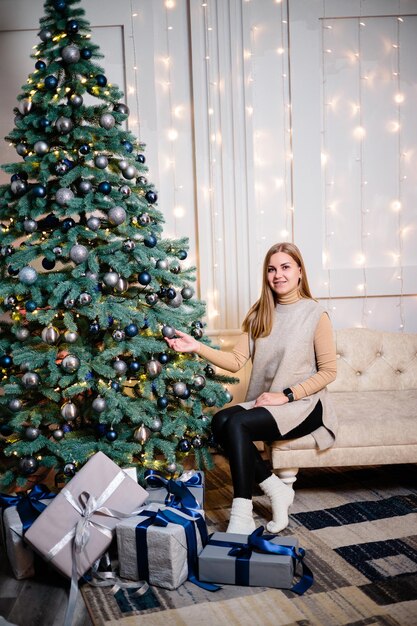 Una hermosa mujer encantadora con un suéter beige se sienta en el piso con el fondo de un árbol de Navidad y decora un árbol de Navidad con juguetes en una espaciosa habitación decorada con guirnaldas.