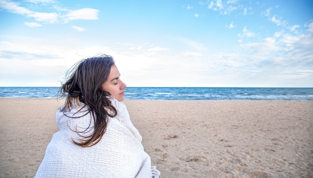 Hermosa mujer envuelta en una manta mira a lo lejos en la playa.