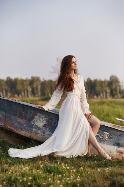 Hermosa mujer esbelta con un vestido blanco largo camina por la mañana cerca del lago. Chica de cabello castaño con cabello largo camina sobre la hierba en el pueblo, cosméticos naturales y maquillaje