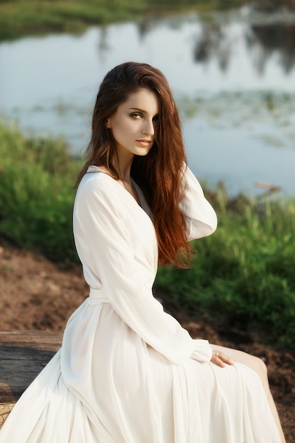 Hermosa mujer esbelta con un vestido blanco largo camina por la mañana cerca del lago.