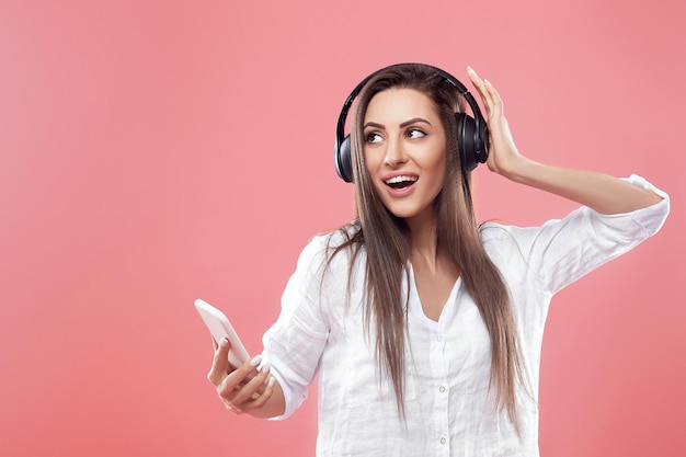 Hermosa mujer escuchando música con auriculares inalámbricos en estudio aislado sobre fondo de color rosa