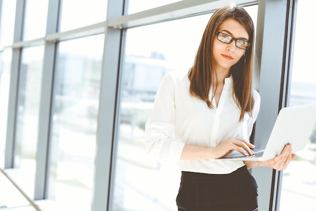 Hermosa mujer especialista con computadora portátil de pie en la oficina moderna y sonriendo encantadoramente Trabajando en la estrategia del plan de análisis de datos de diseño Concepto de personas de negocios