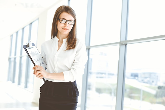 Hermosa mujer especialista con portapapeles de pie en la oficina moderna y sonriendo encantadoramente Trabajando en la estrategia del plan de análisis de datos de diseño Concepto de personas de negocios