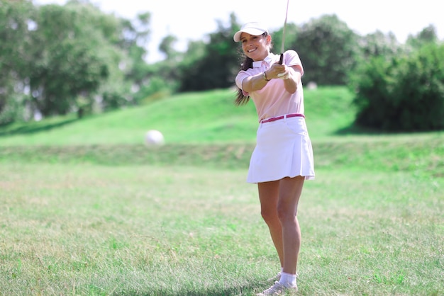 Hermosa mujer en falda blanca, gorra y camiseta rosa jugar al golf en césped verde