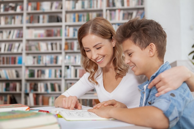 Hermosa mujer feliz abrazando a su hijo pequeño, ayudándolo con el proyecto escolar en la biblioteca