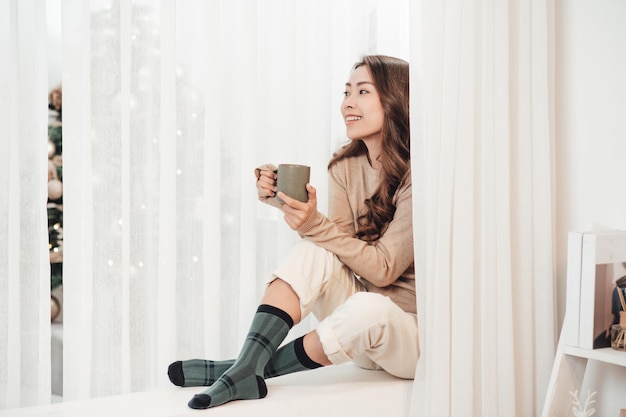 Hermosa mujer feliz bebiendo café caliente sentado en el alféizar de la ventana en casa decorada en Navidad Concepto de vacaciones