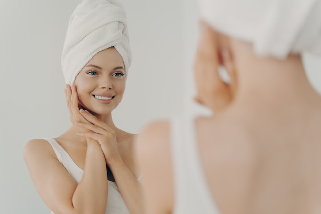 Hermosa mujer feliz después de aplicar maquillaje mirando en el espejo y sonriendo mientras está de pie en el baño, bastante joven con una toalla blanca en la cabeza tocando suavemente la cara. Concepto de belleza y cuidado de la piel.