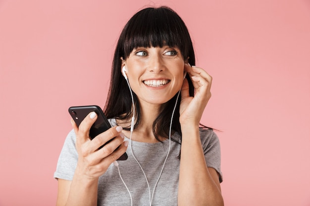 una hermosa mujer feliz emocionada increíble posando aislada sobre la pared de la pared de color rosa claro con teléfono móvil escuchando música con auriculares.