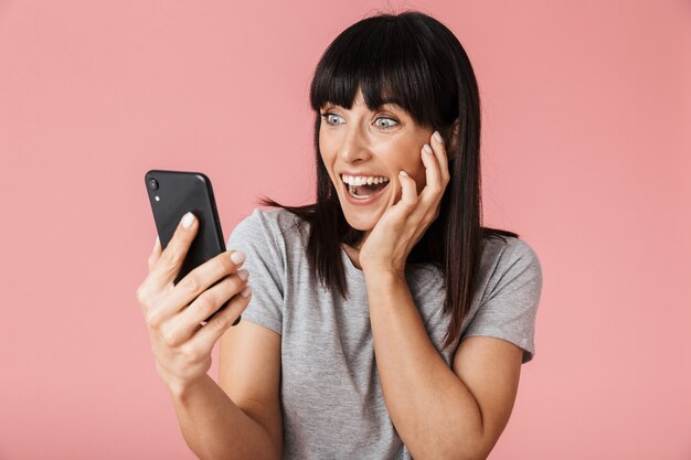 una hermosa mujer feliz emocionada increíble posando aislada sobre la pared de la pared de color rosa claro mediante teléfono móvil.