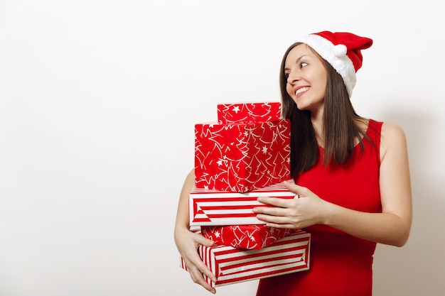 Hermosa mujer feliz joven caucásica con sonrisa encantadora vestida con vestido rojo y sombrero de Navidad con cajas de regalo sobre fondo blanco. Niña de Santa con presente aislado. Concepto de vacaciones de año nuevo 2018