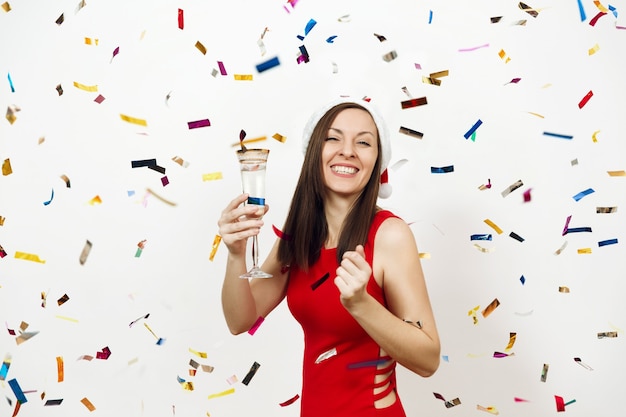 Hermosa mujer feliz joven caucásica con sonrisa encantadora en vestido rojo y sombrero de Navidad de pie con copa de champán sobre fondo blanco y confeti. Niña de Santa aislada. Vacaciones de año nuevo 2018.