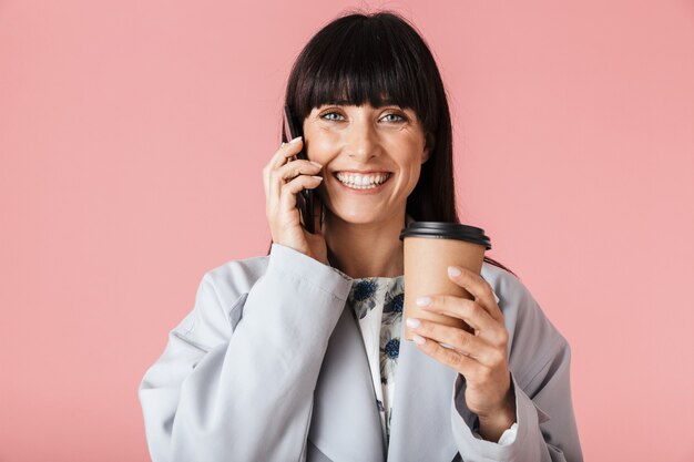 una hermosa mujer feliz posando aislada sobre la pared de la pared de color rosa claro hablando por teléfono móvil con café.