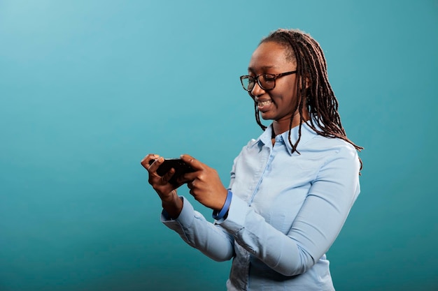 Hermosa mujer feliz con smartphone con pantalla táctil navegando por páginas web de Internet mientras está de pie sobre fondo azul. Seguro joven adulto alegre con teléfono celular moderno viendo contenido de video.
