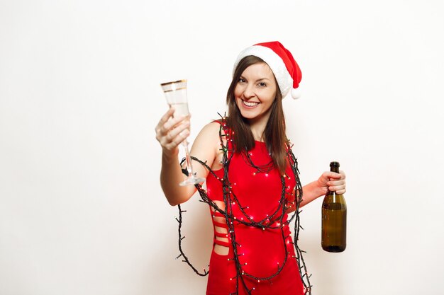 Hermosa mujer feliz sonriente joven encantadora en vestido rojo, sombrero de Navidad, farolillos de guirnalda de pie con vidrio y botella de champán sobre fondo blanco. Niña de Santa aislada. Vacaciones de año nuevo 2018.