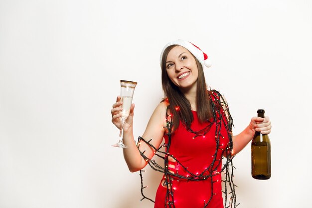 Hermosa mujer feliz sonriente joven encantadora en vestido rojo, sombrero de Navidad, farolillos de guirnalda de pie con vidrio y botella de champán sobre fondo blanco. Niña de Santa aislada. Vacaciones de año nuevo 2018.