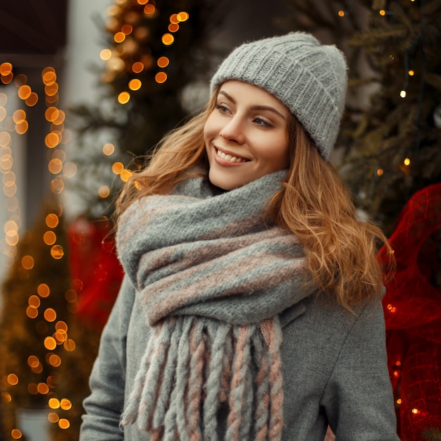 Hermosa mujer feliz con una sonrisa en un abrigo gris sombrero y bufanda de punto de moda cerca de un árbol de Navidad y luces en la ciudad