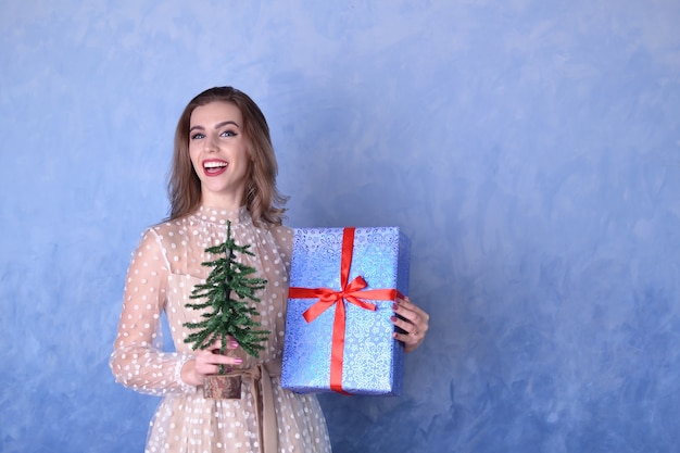 Hermosa mujer feliz sosteniendo un pequeño árbol de Navidad y un regalo azul con una cinta roja en sus manos